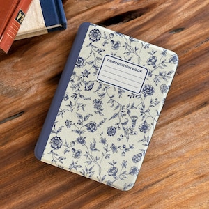 Może przedstawiać: Notatnik z kwiatowym wzorem. Okładka ma kremowe tło z niebieskimi kwiatowymi wzorami i niebieski grzbiet. Etykieta brzmi "COMPOSITION BOOK". Notatnik leży na drewnianej powierzchni.