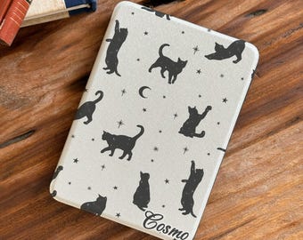 Pode incluir: Uma capa retangular para leitor de e-books com fundo creme claro, apresentando um padrão repetitivo de silhuetas pretas de gatos em várias poses, pequenas estrelas e luas crescentes. O nome Cosmo está impresso em letra preta no canto inferior direito.