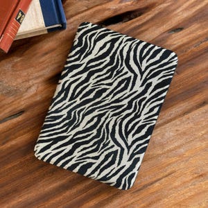 May include: A rectangular, black and white zebra print case. The case is lying on a wooden surface. The zebra print pattern is composed of black stripes on a cream-colored background. The case appears to be a cover for a tablet or notebook.