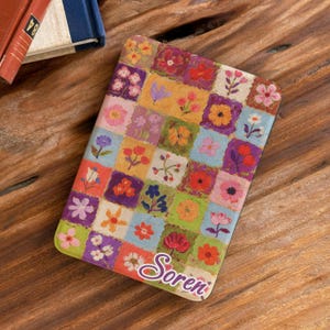Puede incluir: Una funda protectora rectangular con un colorido patrón de cuadrícula de varias pequeñas ilustraciones florales en tonos rojo, amarillo, azul, verde y morado. El nombre Soren está impreso en una tipografía cursiva morada en la esquina inferior derecha.