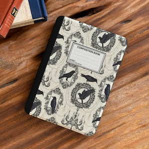May include: Composition book with a cream cover, patterned with black crows in decorative frames and chandeliers. The cover reads "COMPOSITION BOOK" and has a blank label for the owner's name.
