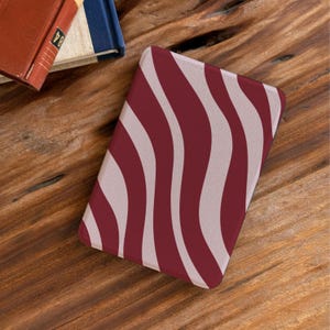 Könnte beinhalten: Eine rechteckige Hülle mit einem wellenförmigen Streifenmuster in Burgunderrot und Off-White. Die Hülle liegt auf einer Holzoberfläche, im Hintergrund ist ein rotes Buch zu sehen.