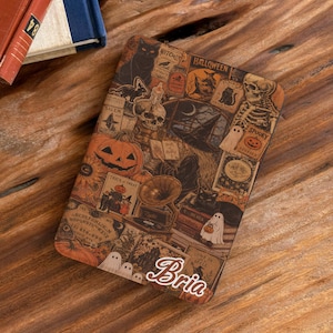 May include: A rectangular e-reader cover featuring a collage of Halloween-themed illustrations including pumpkins, skulls, black cats, ghosts, and a gramophone. The name Bria is printed in a white cursive font at the bottom right corner.