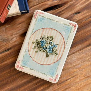 May include: Rectangular tin with a floral design. The center features a bouquet of blue flowers within a pink-striped circle. The border has a light blue background with pink floral accents. The tin is on a wooden surface.