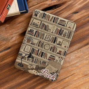 May include: A rectangular e-reader case featuring a printed illustration of a multi-tiered bookshelf filled with books, a desk lamp, a clock, and a cozy chair. The text "Stay Curious," "Collect beautiful moments," "Just one more chapter," and "Home is where the books are" is visible.