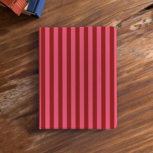 Könnte beinhalten: Ein rechteckiges Notizbuch mit vertikalem Streifenmuster in Rosa- und Rottönen. Das Notizbuch liegt auf einer Holzoberfläche, mit der Ecke von zwei Büchern oben links.