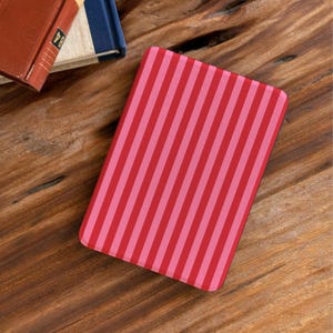 Puede incluir: Un cuaderno rectangular con rayas rosas y rojas descansa sobre una superficie de madera. El lomo de un libro rojo y un libro azul son visibles en el fondo. La cubierta del cuaderno presenta rayas verticales.