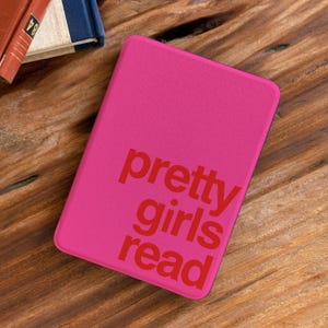 May include: A rectangular e-reader case in a vibrant hot pink color. The front surface features the text "pretty girls read" printed in a bold, dark red font. The case has rounded corners and a smooth, matte-textured finish.
