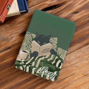 May include: A dark green e-reader case featuring a black cat reading a book titled COMMENT DOMESTIQUER LES HUMAINS, with a cup of coffee and striped bedding. The name Rory is printed in white cursive script at the bottom of the cover.