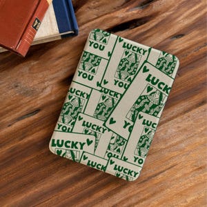 May include: A rectangular, cream-coloured phone case with a repeating pattern of playing card designs in green. The cards feature a queen illustration, the word "LUCKY," and the word "YOU."