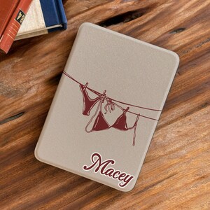 May include: A rectangular beige e-reader case featuring a dark red line art illustration of a bikini and underwear hanging on a clothesline. The name Macey is printed in a dark red cursive script font below the illustration on the front cover.
