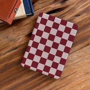 Może przedstawiać: Prostokątne etui z bordowo-białym wzorem w szachownicę. Etui leży na drewnianej powierzchni, z rogiem czerwonej książki i niebieskiej książki w tle.