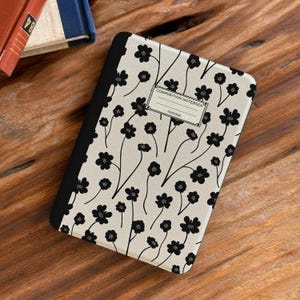 Peut inclure: Un cahier de composition avec un motif floral. La couverture présente un fond crème avec des fleurs et des tiges noires. La tranche est noire et le cahier porte une étiquette indiquant "COMPOSITION NOTEBOOK".