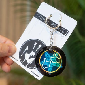 May include: A black and iridescent keyring with a silver chain and ring. The keyring features a circular design with two thumbs-up symbols and a star. The card behind the keyring reads "CHIRAL PRINTS" and has a handprint logo.