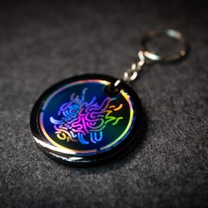 K&ouml;nnte beinhalten: Ein runder, schwarzer Schl&uuml;sselanh&auml;nger mit einem farbenfrohen, irisierenden Design. Das Design zeigt ein stilisiertes, abstraktes Muster in Blau-, Lila- und Gelbt&ouml;nen. Der Schl&uuml;sselanh&auml;nger hat eine silberne Kette und einen Ring.