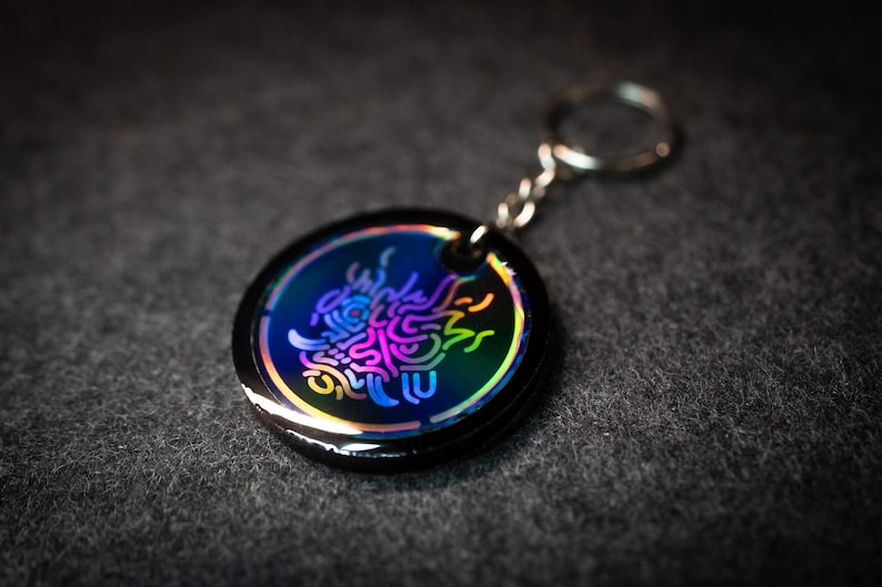 K&ouml;nnte beinhalten: Ein runder, schwarzer Schl&uuml;sselanh&auml;nger mit einem farbenfrohen, irisierenden Design. Das Design zeigt ein stilisiertes, abstraktes Muster in Blau-, Lila- und Gelbt&ouml;nen. Der Schl&uuml;sselanh&auml;nger hat eine silberne Kette und einen Ring.