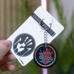 K&ouml;nnte beinhalten: Ein schwarzer Schl&uuml;sselanh&auml;nger mit einem farbenfrohen Design und einer silbernen Kette. Der Schl&uuml;sselanh&auml;nger hat einen runden Anh&auml;nger mit Neon-Design. Die beigef&uuml;gte Karte tr&auml;gt den Text "CHIRAL PRINTS" und einen schwarz-wei&szlig;en Handabdruck.