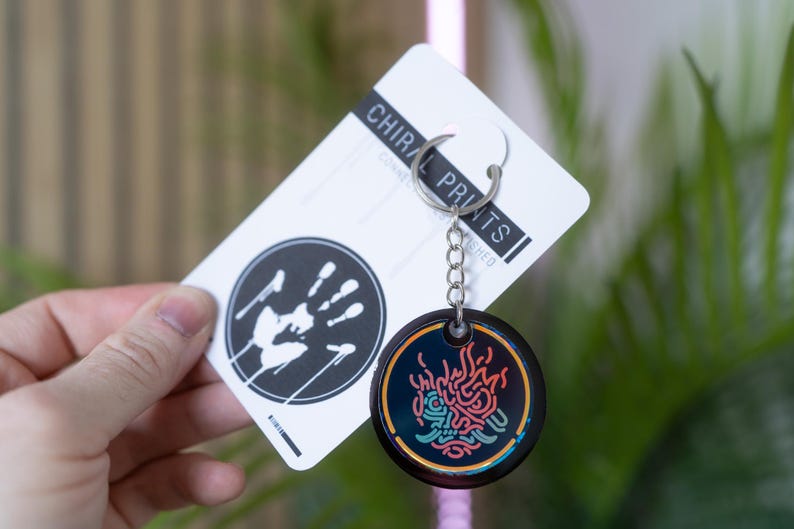 K&ouml;nnte beinhalten: Ein schwarzer Schl&uuml;sselanh&auml;nger mit einem farbenfrohen Design und einer silbernen Kette. Der Schl&uuml;sselanh&auml;nger hat einen runden Anh&auml;nger mit Neon-Design. Die beigef&uuml;gte Karte tr&auml;gt den Text "CHIRAL PRINTS" und einen schwarz-wei&szlig;en Handabdruck.