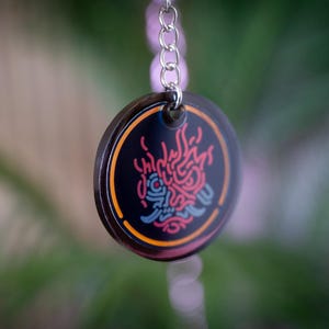 K&ouml;nnte beinhalten: Ein runder, schwarzer Schl&uuml;sselanh&auml;nger-Anh&auml;nger mit farbenfrohem Design. Der Anh&auml;nger hat ein rotes, blaues und orangefarbenes Design auf schwarzem Hintergrund. Der Anh&auml;nger ist an einer silbernen Kette befestigt. Der Hintergrund ist verschwommen gr&uuml;n und braun.