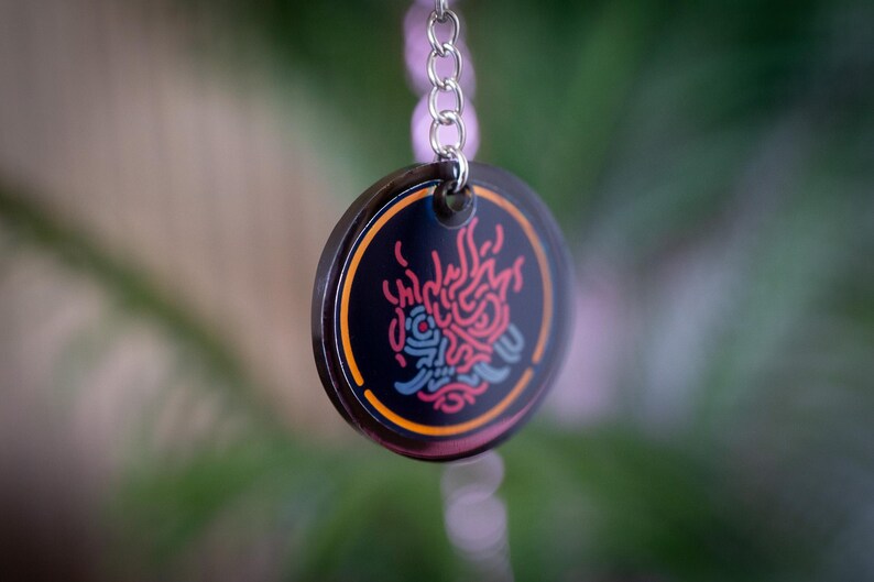 K&ouml;nnte beinhalten: Ein runder, schwarzer Schl&uuml;sselanh&auml;nger-Anh&auml;nger mit farbenfrohem Design. Der Anh&auml;nger hat ein rotes, blaues und orangefarbenes Design auf schwarzem Hintergrund. Der Anh&auml;nger ist an einer silbernen Kette befestigt. Der Hintergrund ist verschwommen gr&uuml;n und braun.