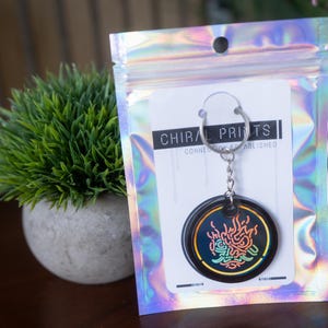 K&ouml;nnte beinhalten: Ein schwarzer Schl&uuml;sselanh&auml;nger mit einem kreisf&ouml;rmigen Design mit einer farbenfrohen, abstrakten Grafik. Der Schl&uuml;sselanh&auml;nger ist an einer Karte in einer holografischen Zip-Tasche befestigt. Der Text "CHIRAL PRINTS" ist auf der Karte zu sehen.