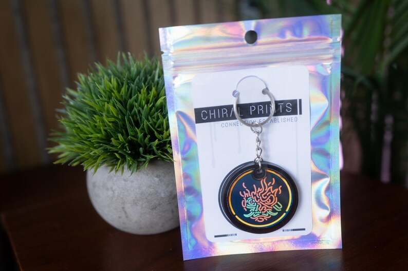K&ouml;nnte beinhalten: Ein schwarzer Schl&uuml;sselanh&auml;nger mit einem kreisf&ouml;rmigen Design mit einer farbenfrohen, abstrakten Grafik. Der Schl&uuml;sselanh&auml;nger ist an einer Karte in einer holografischen Zip-Tasche befestigt. Der Text "CHIRAL PRINTS" ist auf der Karte zu sehen.