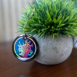 K&ouml;nnte beinhalten: Ein runder, schwarzer Schl&uuml;sselanh&auml;nger mit einer silbernen Kette. Der Schl&uuml;sselanh&auml;nger zeigt ein farbenfrohes, stilisiertes Design mit blauen, rosa und gelben Farbt&ouml;nen. Der Schl&uuml;sselanh&auml;nger wird neben einem kleinen, runden, grauen Topf mit gr&uuml;nen Pflanzen pr&auml;sentiert.