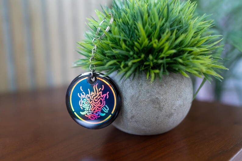 K&ouml;nnte beinhalten: Ein runder, schwarzer Schl&uuml;sselanh&auml;nger mit einer silbernen Kette. Der Schl&uuml;sselanh&auml;nger zeigt ein farbenfrohes, stilisiertes Design mit blauen, rosa und gelben Farbt&ouml;nen. Der Schl&uuml;sselanh&auml;nger wird neben einem kleinen, runden, grauen Topf mit gr&uuml;nen Pflanzen pr&auml;sentiert.