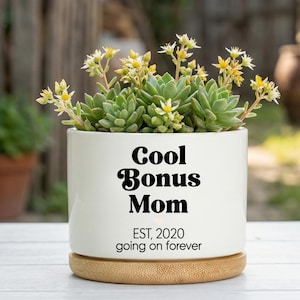 May include: A white ceramic planter with the text "Cool Bonus Mom" and "EST. 2020 going on forever" in black. The planter holds a succulent plant with green leaves and small yellow flowers, sitting on a wooden base.