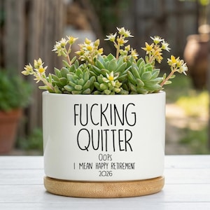 May include: A white ceramic planter with the text "FUCKING QUITTER" and "OOPS I MEAN HAPPY RETIREMENT 2026" in black. The planter holds a succulent plant with green leaves and small yellow flowers. It sits on a wooden base.