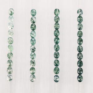 May include: A collection of oval-cut moss agate gemstones, each displaying unique patterns of green and white inclusions. The gemstones are arranged in vertical rows against a white background, showcasing their natural beauty and variations.