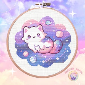 Puede incluir: Bordado de punto de cruz en un aro de madera que presenta un gato blanco con detalles rosas, sobre un diseño de nube con planetas y estrellas. El fondo es una gradación de rosa, morado y azul, con el texto "xstitch_verse".