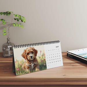 May include: A desk calendar featuring a watercolor illustration of a brown and white puppy in a field of pink flowers. The calendar is open to May, with the month's name and dates visible. The calendar is on a wooden surface.