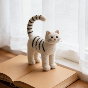 May include: A white cat ornament with black stripes and a curved tail is positioned on an open book. The cat has detailed features, including whiskers. The book rests on a wooden surface, near a window with white curtains. The cat is approximately 15 cm tall.