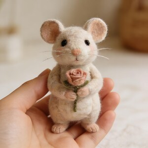 Puede incluir: Una figura de rat&oacute;n artesanal de fieltro blanco roto sosteniendo una rosa rosa. El rat&oacute;n tiene orejas grandes y redondas, ojos negros y bigotes delicados. La figura se sostiene en una mano, mostrando los intrincados detalles del trabajo en fieltro.