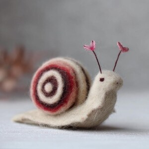 Puede incluir: Una figura de caracol de fieltro hecha a mano. La concha del caracol presenta rayas concéntricas crema, burdeos y marrón. Tiene un cuerpo color crema y dos antenas rosas. La figura es un objeto decorativo caprichoso.