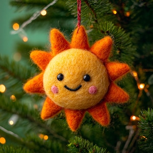 Może przedstawiać: Ręcznie robiona filcowa ozdoba choinkowa w kształcie słońca. Słońce ma uśmiechniętą twarz, czarne oczy i różowe policzki. Posiada pomarańczowe promienie i żółty środek. Wisi na czerwonym sznurku.