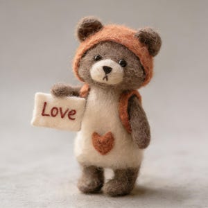 Puede incluir: Un oso de peluche de fieltro hecho a mano con una chaqueta con capucha y mochila naranja, sosteniendo un pequeño letrero que dice "Love". El oso tiene un cuerpo blanco con un corazón marrón y detalles marrones.