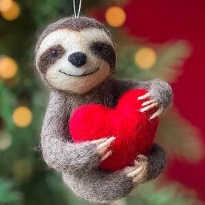 May include: A handmade sloth ornament made of gray felt, holding a bright red heart. The sloth has a smiling face with brown markings and a white string for hanging. The ornament is set against a blurred background of green and red.