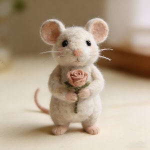 Puede incluir: Una figura de rat&oacute;n de fieltro blanco hecha a mano que sostiene una rosa rosa. El rat&oacute;n tiene orejas grandes y redondeadas, ojos negros y bigotes delicados. Sus patas y los p&eacute;talos de la rosa son de color rosa claro. La cola del rat&oacute;n es de color rosa p&aacute;lido.