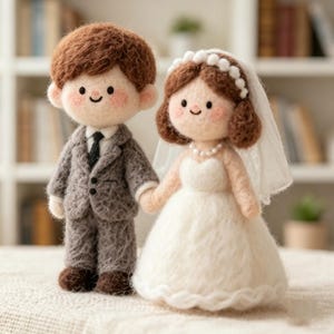Puede incluir: Dos muñecos de lana fieltrada que representan a una pareja. La figura de la izquierda tiene el pelo castaño y viste un traje gris con corbata negra. La figura de la derecha tiene el pelo castaño, velo blanco con diadema de perlas y vestido blanco.