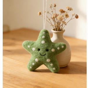 Puede incluir: Adorno de fieltro en forma de estrella de mar verde con lunares blancos y una cara sonriente. Cuelga de un hilo blanco. Un pequeño jarrón con flores secas se encuentra en el fondo.