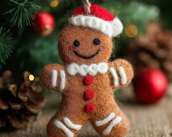 以下が含まれることがあります： フェルト製のジンジャーブレッドマンのクリスマスオーナメント。茶色のオーナメントには白いアイシングのディテール、赤いサンタ帽、赤いボタンが付いています。天然の麻ひもで吊るされています。背景にはクリスマスツリーと赤いオーナメントがあります。