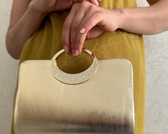 May include: A gold clutch with a circular handle adorned with sparkling rhinestones. The rectangular bag has a textured surface and a metallic sheen. The handle is held by a person wearing a yellow dress.