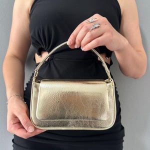 Könnte beinhalten: Eine goldfarbene Handtasche mit rechteckiger Form und Klappenverschluss. Die Tasche hat eine strukturierte Oberfläche und einen dünnen, goldenen Griff. Die Tasche wird von einer Person gehalten, die ein schwarzes Kleid und silberne Ringe trägt.