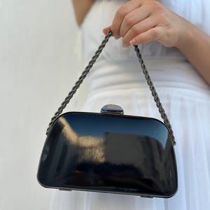 Puede incluir: Un bolso de mano negro brillante con un cierre plateado y una cadena trenzada. El bolso rectangular tiene bordes redondeados y refleja la luz. Un accesorio elegante para eventos.