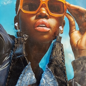 May include: Black leather and denim earrings. The earrings are a unique design, with a triangular shape. The top portion is black leather, and the bottom is denim. The earrings are hanging from a clear earring post.