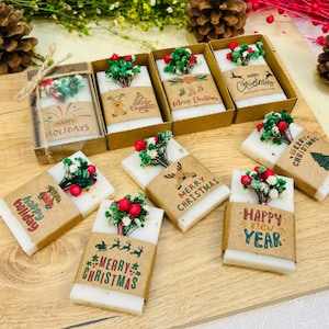 May include: A collection of handmade soaps with festive decorations. Each soap is adorned with a small bouquet of red berries and greenery, and wrapped with a brown paper label. The labels feature holiday greetings like "Happy Holidays," "Merry Christmas," and "Happy New Year."