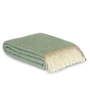 Peut inclure: Une couverture pliée, vert sauge, avec un motif en losanges. La couverture a une bordure beige et une frange crème sur un bord. La couverture douce est probablement en laine ou dans une matière similaire.