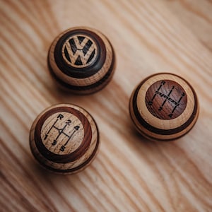 Puede incluir: Tres pomos de cambio de madera con veta natural. Uno presenta el logotipo de Volkswagen, los otros muestran patrones de cambio de marchas. Los pomos son de madera clara y oscura alternada, sobre una superficie de madera.